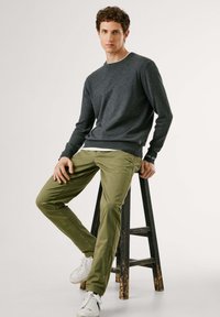 Pull en tricot gris foncé avec un col rond, pantalon vert slim et baskets blanches. Modèle assis sur un tabouret en bois noir.