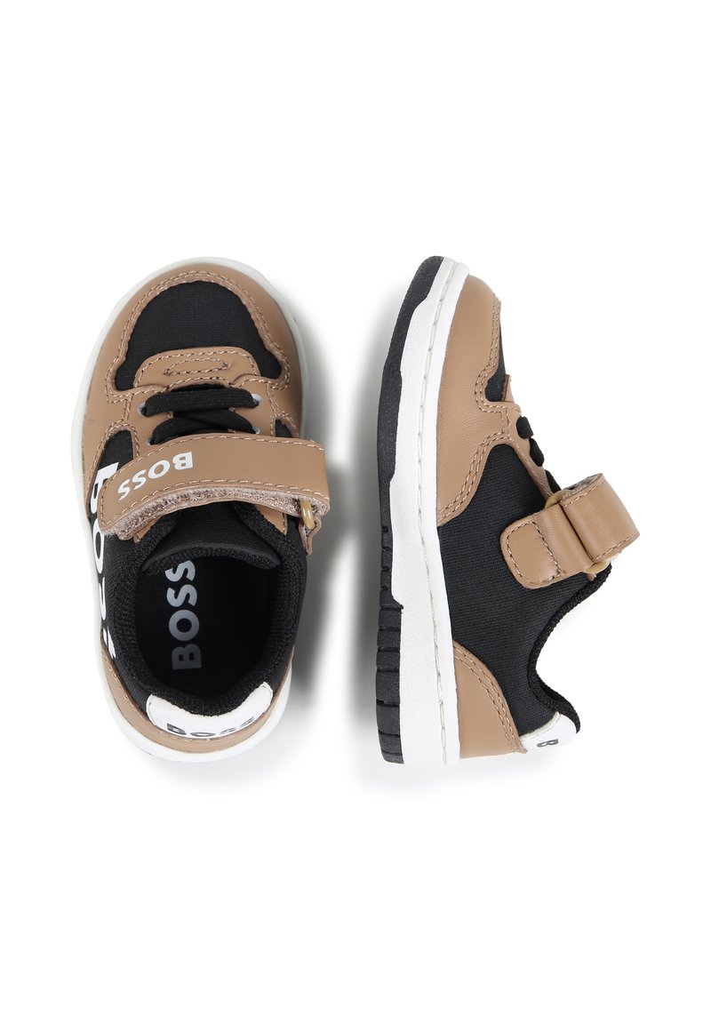 Zapatillas en tonos marrones y negras con una tira de velcro, acentos blancos, material de nailon, suela acolchada y detalles del logo en el lateral y el talón.