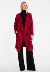 Rot-karierter Wickelcardigan aus weichem Material, mit Fransenabschluss, offener Vorderseite und zwei Taschen. Kombiniert mit schwarzen weit geschnittenen Hosen.