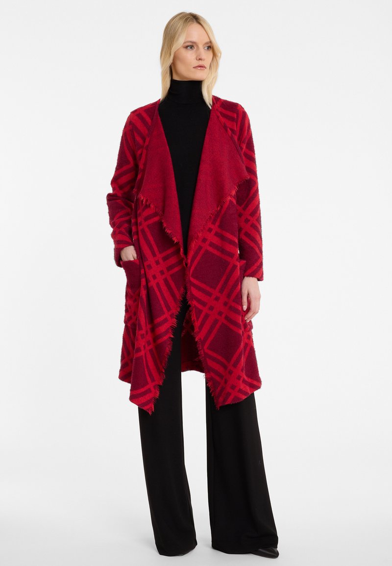 Rot-karierter Wickelcardigan aus weichem Material, mit Fransenabschluss, offener Vorderseite und zwei Taschen. Kombiniert mit schwarzen weit geschnittenen Hosen.