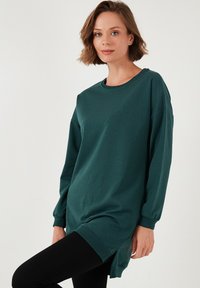 Túlméretezett zöld pulóver hosszú ujjal, kerek nyakkivágással, oldalsó hasítékokkal és bordás mandzsettával. Puha pamut anyagból készült.