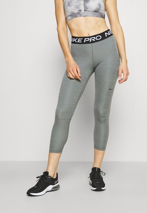 Kobieta ubrana w szare legginsy Nike Pro, czarno-białe buty sportowe Nike oraz szary, wzorzysty biustonosz sportowy, stojąca na jednolitym tle.