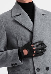 Šedý vlněný kabát s klopami s výřezem. Černé kožené rukavice s odkrytými prsty a knoflíkovým detailem, držené proti kabátu.