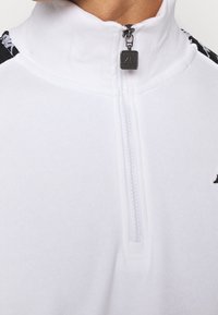 Bílá mikina na zip s vysokým límcem, ozdobená černým vzorovaným motivem na ramenou a stříbrným zipem s logem.