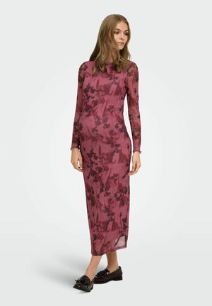 Mujer de pie mirando de lado, lleva un vestido midi ajustado de manga larga con un patrón floral oscuro y mocasines oscuros.