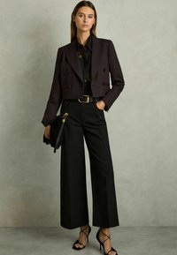 Zwarte dubbelgeknoopte cropped blazer over een zwarte bloes met bloemenmotief, gecombineerd met hoge, zwarte wijde broekspijpen en zwarte sandaaltjes met bandjes.