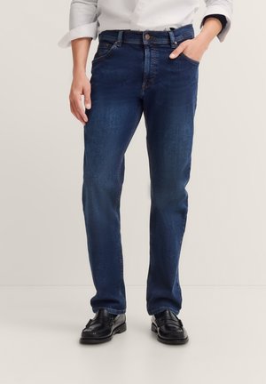 Man wearing dark blue jeans, white rolled-up shirt, and black leather loafers, standing with one hand in pocket against plain background.