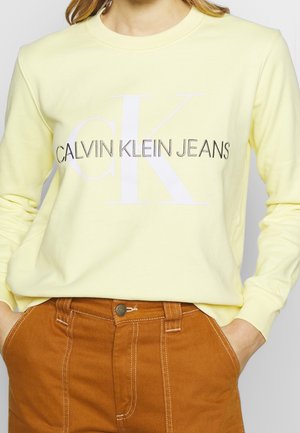 Osoba nosząca bladożółtą bluzę Calvin Klein Jeans i wysokowysokie brązowe spodnie z rękami w kieszeniach.