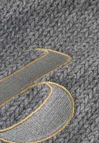 Detailný záber na štruktúrovanú sivú látku s veľkým vyšívaným štýlovým písmenom "J" orámovaným svetlohnedou niťou.