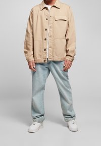 Beige bomullsjacka med framfickor och knäppning framtill, kombinerad med ljusblå baggyjeans och vita sneakers. Texturerad, avslappnad design.