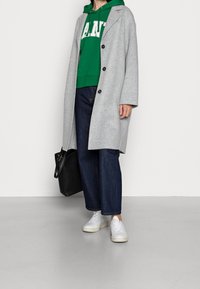 Cappotto grigio in lana con bottoni neri indossato sopra una felpa con cappuccio verde, abbinato a pantaloni larghi in denim scuro e sneakers bianche. Borsa nera inclusa.
