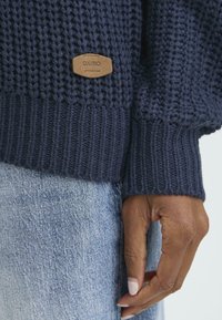 Mørkeblå strikket sweater med struktureret vævning og ribbede manchetter, prydet med en lysebrun læderetsikette med inskriptionen "OXMO." Sammen med lyse denimbukser.