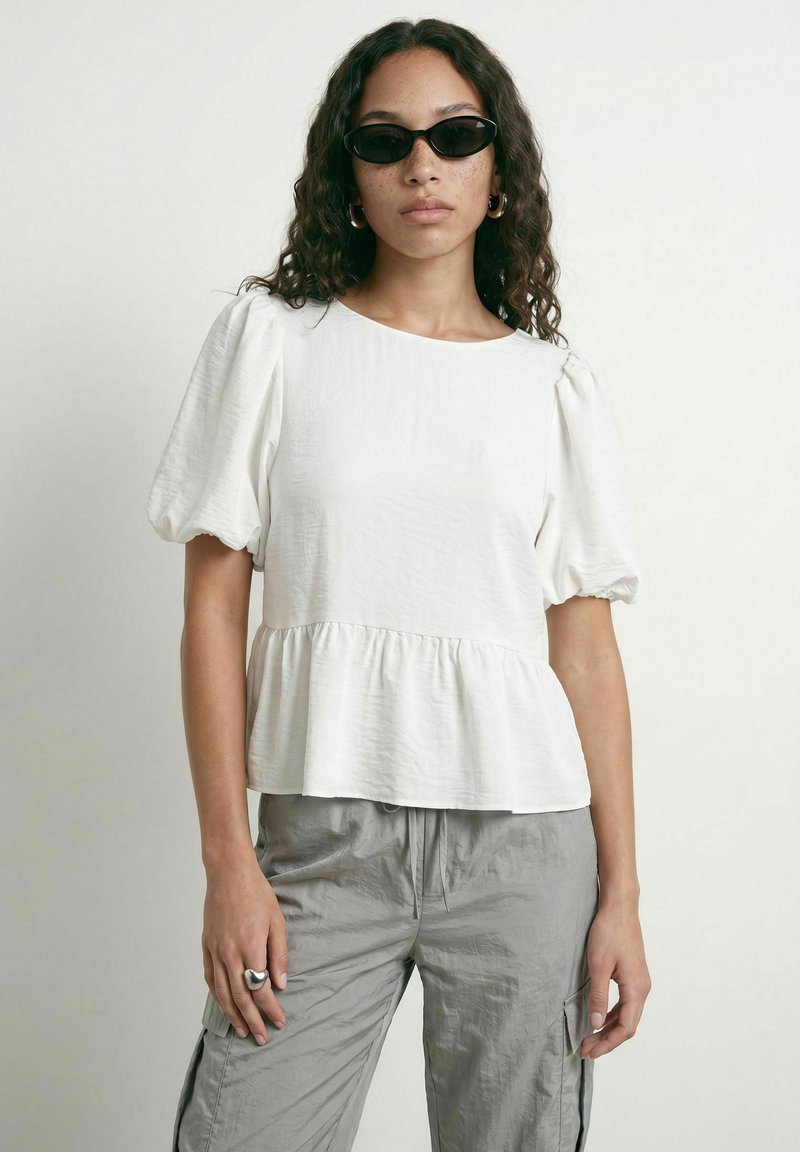 Femme aux cheveux bouclés portant des lunettes de soleil noires, un chemisier blanc à manches bouffantes, un pantalon cargo gris, des boucles d’oreilles anneaux et une bague en argent à la main gauche.