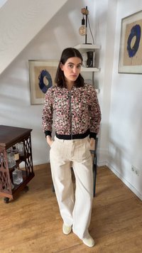 Femme debout à l'intérieur portant une veste zippée florale, un pantalon large crème et des chaussures beiges dans une pièce avec un sol en bois et des œuvres encadrées.