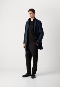 Cappotto impermeabile blu navy con colletto e chiusura a bottoni, abbinato a un maglione nero e pantaloni, completato da scarpe nere.