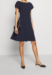 Námorně modré šaty s krátkými balónovými rukávy, silueta ve tvaru A a délka ke kolenům. Nosené s černými botami na podpatku typu slingback a malou kabelkou.