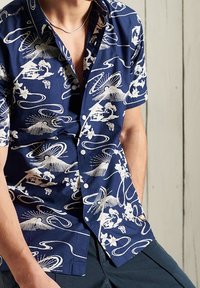 Camisa de botones de manga corta en color azul marino con impresiones gráficas blancas de montañas, olas y patrones florales en toda la tela.