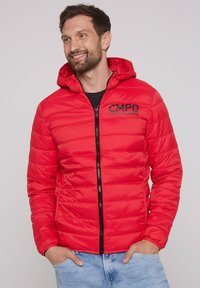 Rote gepolsterte Jacke mit Kapuze; gesteppter Design; schwarzer Reißverschluss und Logo auf der Brust; lange Ärmel. Getragen mit einem blauen Hemd und hellen Jeans.