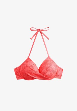 Kvinde med langt krøllet hår iført en koralrød tekstureret bikini, stående mod en lysefarvet væg i naturligt sollys.
