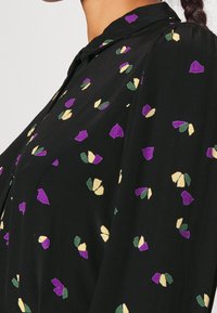 Tissu noir avec un petit motif floral violet, jaune et vert sur un vêtement à col porté par une personne.