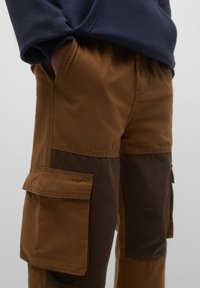 Mand iført brune og mørkebrun to-tone cargobukser med hænderne i lommerne, kombineret med en marineblå sweatshirt.
