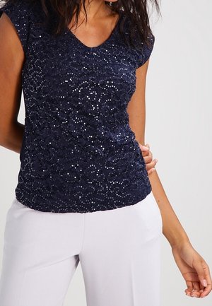 Kobieta w granatowym krótkim topie z cekinami, w połączeniu z białymi spodniami z wysokim stanem, na jednolitym tle.