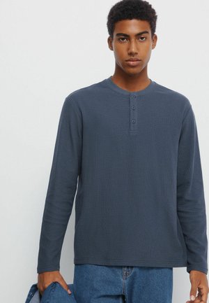 Marineblå langærmet henley-skjorte med tre-knaps placket, ribbet tekstur, rund halsudskæring og lidt afslappet pasform, iført med blå jeans.