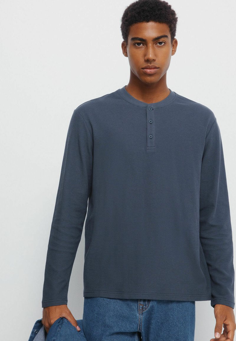 Marineblaues Langarm-Henley-Shirt mit Drei-Knopf-Leiste, gerippter Textur, rundem Halsausschnitt und leicht lockerem Schnitt, getragen mit blauen Jeans.