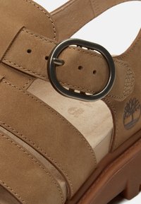 Hnědá kožená sandála s kulatou kovovou sponou, nastavitelným páskem, polstrovanou stélkou a logem stromu Timberland na boku.