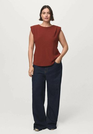 Woman with short dark hair wearing a sleeveless rust-colored top and dark wide-leg jeans, standing with one hand in her pocket.