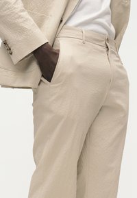 Άτομο με σκούρο δέρμα που φοράει υφασμάτινο μπεζ κοστούμι με το ένα χέρι στην τσέπη, με λευκό πουκάμισο να φαίνεται κάτω από το σακάκι.