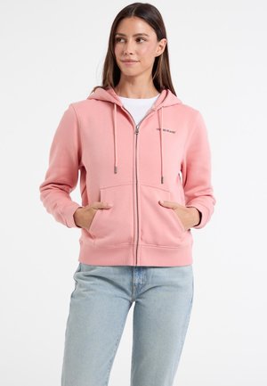 Rosa zip-up hættetrøje lavet af blødt stof, med en kængurulomme, snoretræk i hætten og logo-detalje på venstre bryst. Sammen med lyse jeans.