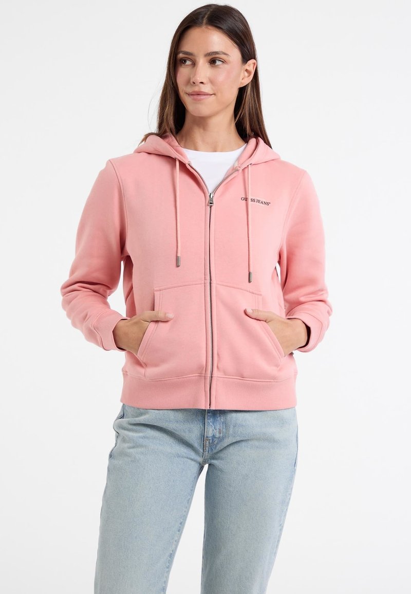 Felpa con zip rosa realizzata in tessuto morbido, dotata di tasca a marsupio, cappuccio con lacci e dettaglio del logo sul lato sinistro del petto. Abbinata a jeans chiari.
