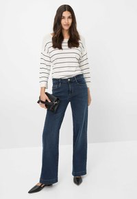 Gestreiftes Oberteil mit langen Ärmeln in Schwarz und Weiß, kombiniert mit dunkelblauen, weit geschnittenen Jeans. Das Model hält eine kleine schwarze Clutch. Spitz zulaufende schwarze Schuhe.