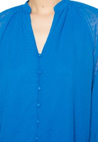 Blusa azul com decote em V e fecho de botão, apresentando um padrão texturizado e mangas transparentes. O material é leve e suave.