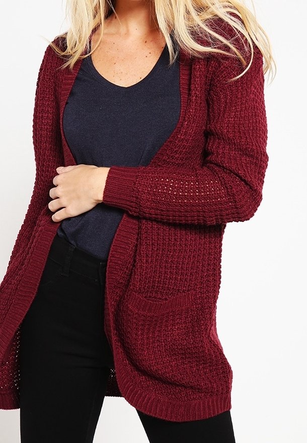 Femme portant un cardigan en tricot rouge foncé sur une chemise bleu marine et un pantalon noir, une main reposant sur sa taille.