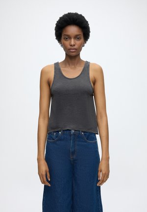 Γυναίκα με κοντά σγουρά μαλλιά, που φοράει ένα αμάνικο σκούρο γκρι τοπ και ψηλόμεσα μπλε τζιν, στέκεται μπροστά σε ένα μονόχρωμο φόντο.