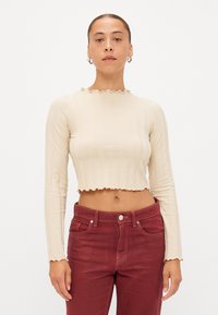 Beige cropped langærmet top med ribbet tekstur og volangkrave, parret med højtaljede maroon jeans.
