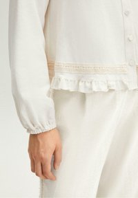 Primer plano de una persona que lleva una blusa de manga larga color crema con detalles de encaje y dobladillo de volantes, combinada con pantalones texturizados color crema.