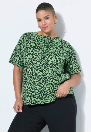 Persoon met kort haar die gouden oorringen draagt en een groene top met luipaardprint, poseert met één hand op de heup tegen een witte achtergrond.