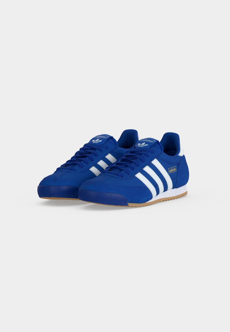 Blauwe Adidas sneakers met witte strepen, witte hielstukjes en zolen van gumrubber, naast elkaar getoond op een effen achtergrond.