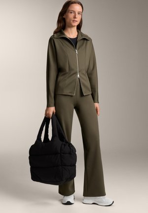 Olajzöld cipzáras dzseki gallérral, hozzá passzoló, fazonra szabott nadrággal. Fekete puffertáska a kézben, fehér sportcipő. Sima felület.