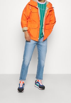 Chaqueta impermeable naranja con forro beige, camiseta a rayas verde, jeans de mezclilla azul claro y zapatillas multicolor con acentos negros.