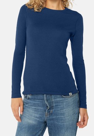 Nainen pukeutuneena istuvaan tummansiniseen pitkähihaseen paitaan ja vaaleansinisiin farkkuihin, seisoo käsivarret rentoina sivuillaan.