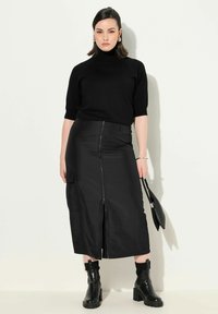 Schwarzer Rollkragenpullover mit kurzen Ärmeln, kombiniert mit einem langen, schwarzen, mit Reißverschluss versehenen Rock und klobigen schwarzen Stiefeletten, während eine schwarze Handtasche gehalten wird.