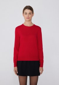 Pull en laine rouge à col rond, à manches longues et texture lisse. Assorti à une jupe noire, créant un look superposé.