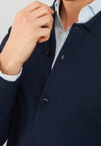 Cardigan blu navy con tessuto testurizzato, dotato di chiusura con bottoni, un piccolo logo ricamato e una camicia azzurro chiaro sovrapposta.