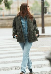 Grøn quiltet jakke over en denimskjorte, parret med lys blå flared jeans og brune sneakers. Outfittet har lommer og en afslappet pasform.