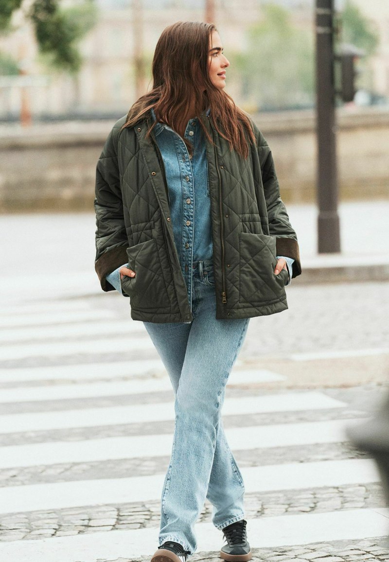 Grøn quiltet jakke over en denimskjorte, parret med lys blå flared jeans og brune sneakers. Outfittet har lommer og en afslappet pasform.