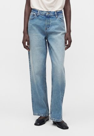 Person iført lyseblå jeans med vide ben og sorte sko med firkantet tå, stående mod en ensfarvet hvid baggrund.